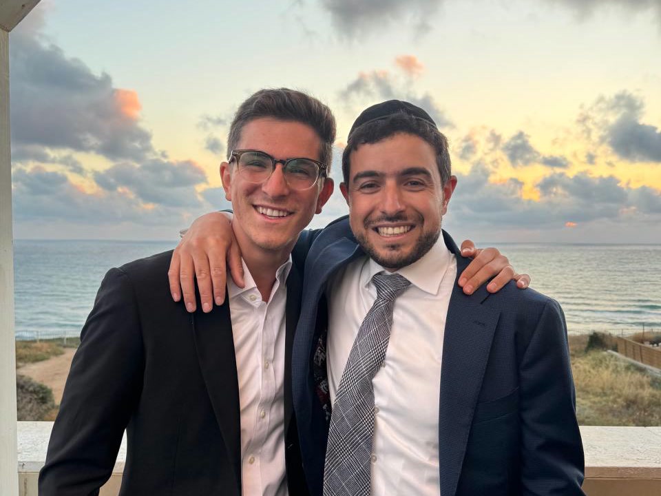 Rabbi Adam Abergel and a young man, arms around each other, on a balcony above the sea at sunset.